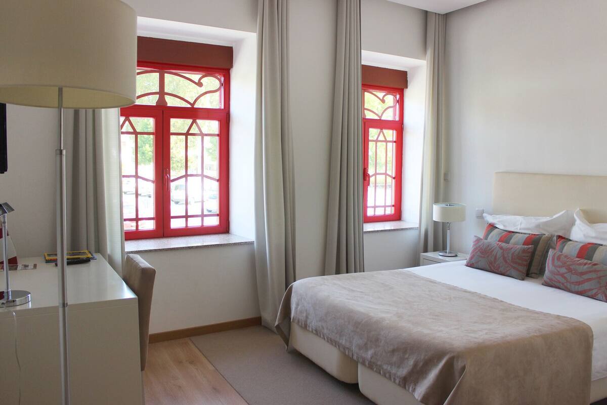 Une chambre d’hôtel avec un lit, un bureau, deux fenêtres à cadre rouge et une lampe.