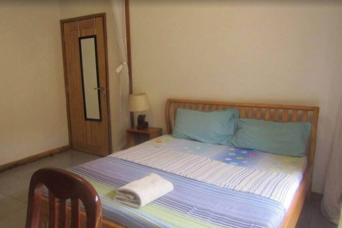 A bedroom with a wooden bed, two pillows, a bedside table with a lamp, a chair, and a door.