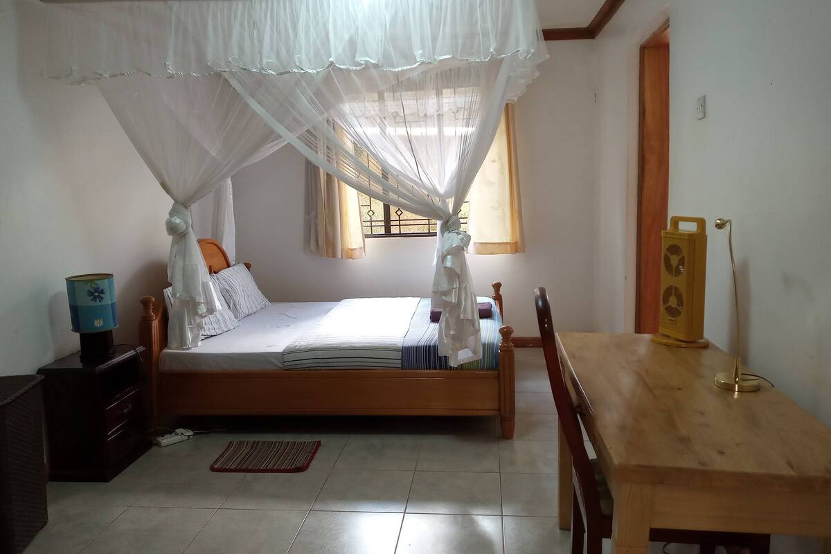 A bedroom with a bed, bedside table, lamp, and a wooden table with a speaker.