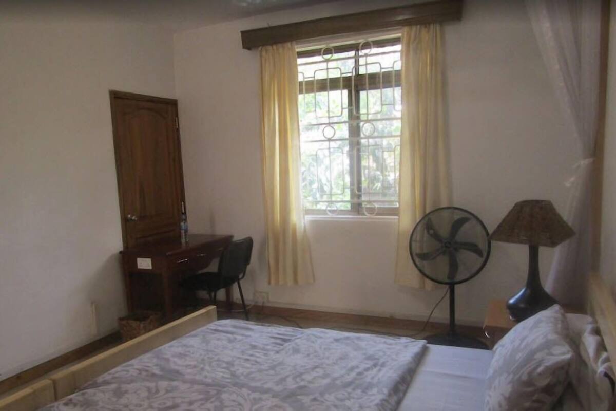 A bedroom with a bed, a desk, a chair, a window with curtains, and a standing fan.
