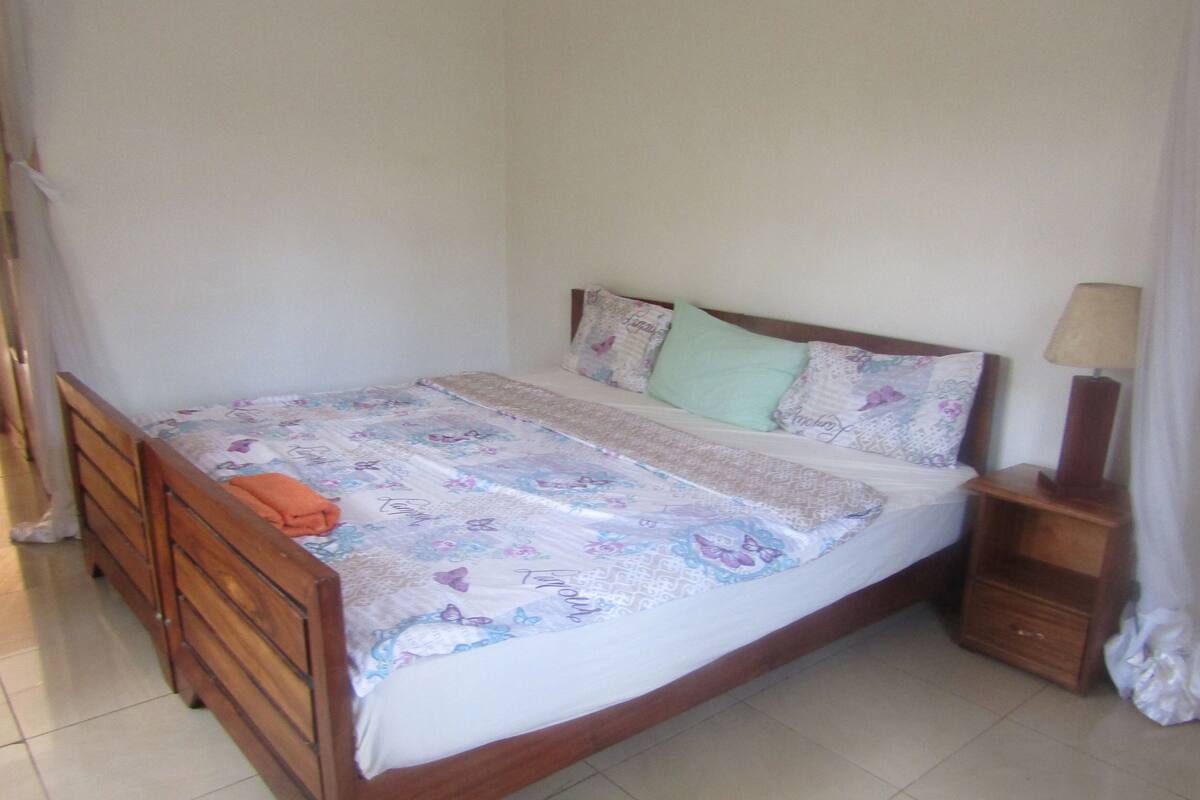 A single bed with a floral bedspread, two pillows, a nightstand with a lamp, and a wooden wardrobe.
