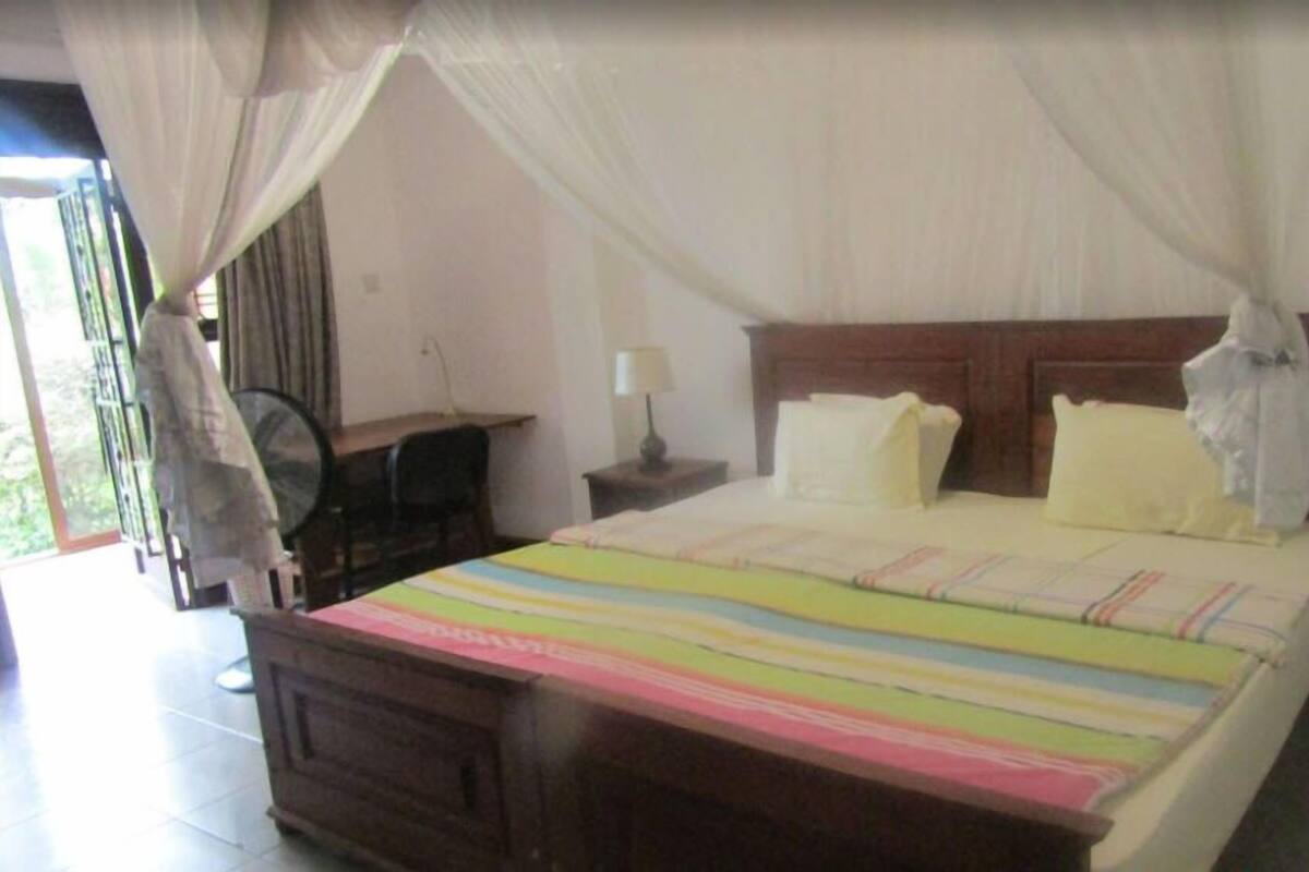 A bedroom with a bed, a nightstand, a bedside table, a lamp, a fan, and a window with curtains.