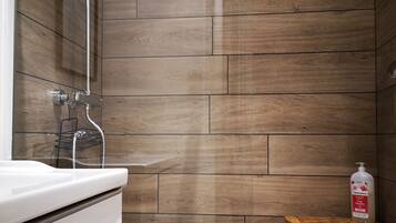 Une salle de bain moderne avec un mur décoré en bois, un lavabo blanc, une douchette à main et un petit tabouret en bois sur lequel est posée une bouteille de lotion.
