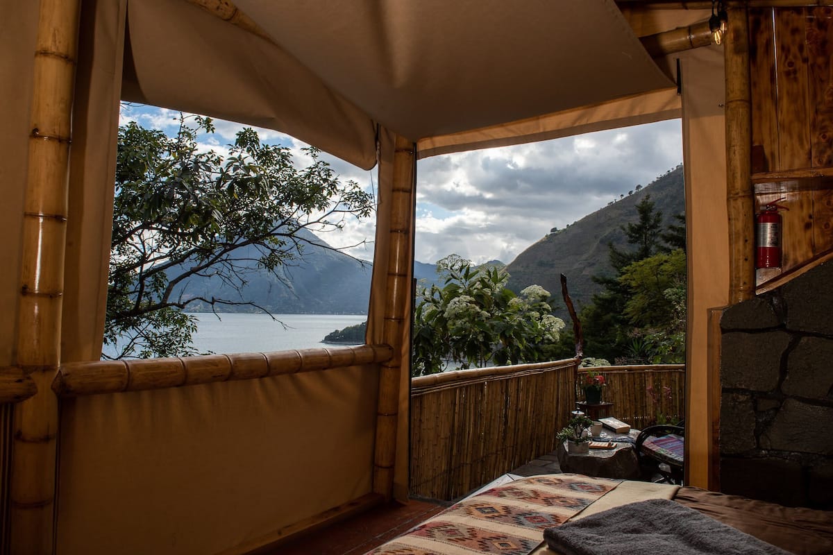 A room with a view of a lake and mountains, bamboo walls, and a stone fireplace.
