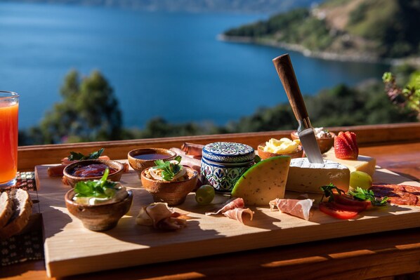 Una tabla de madera con diversos quesos, carnes y frutas, frente a una vista panorámica de un lago.