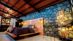 A bedroom with a stone wall, a wooden ceiling, a bed with pillows, a chair, and a shelf with items. - El Picnic Atitlan (Santa Cruz La Laguna)