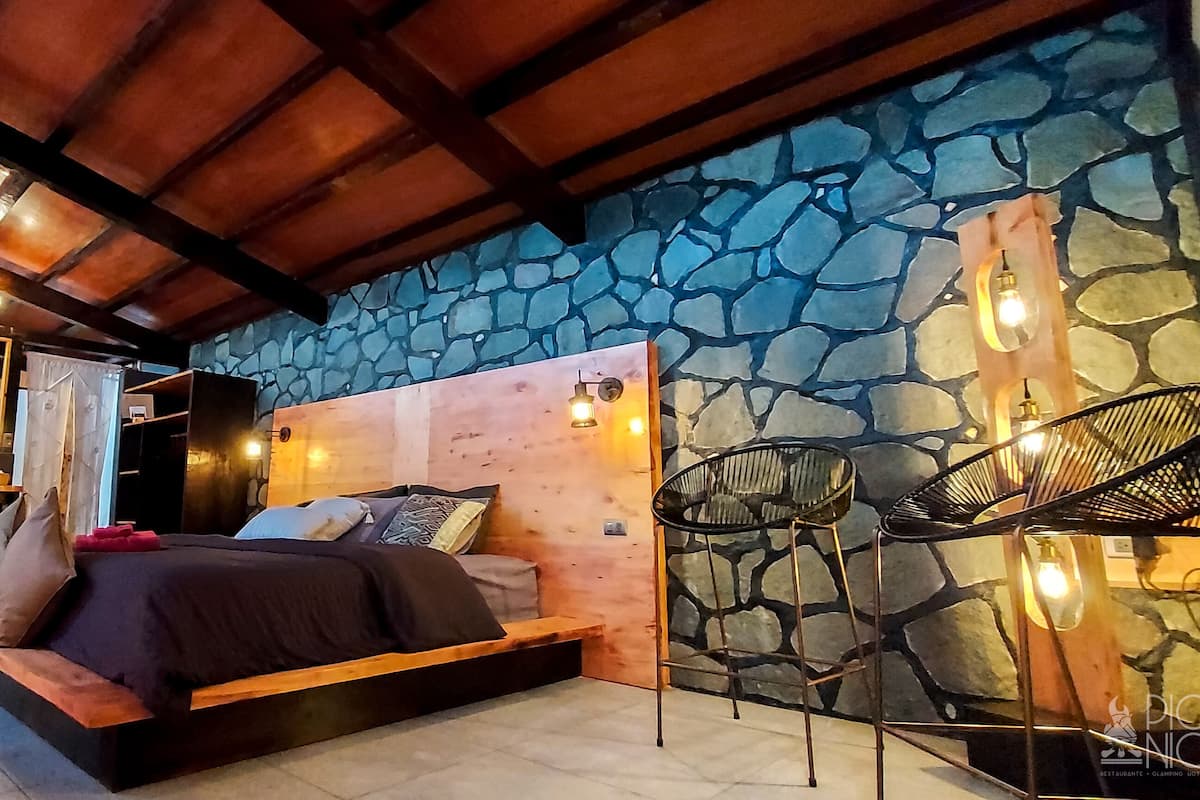 A bedroom with a stone wall, a wooden ceiling, a bed with pillows, a chair, and a shelf with items.