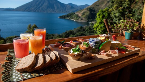A table with a variety of food and drinks, including bread, cheese, and fruit, with a scenic view of a lake and mountains in the background. - El Picnic Atitlan (Santa Cruz La Laguna)
