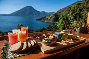 Una mesa con una variedad de comida y bebidas, incluyendo pan, queso y fruta, con una vista panorámica de un lago y montañas al fondo.
