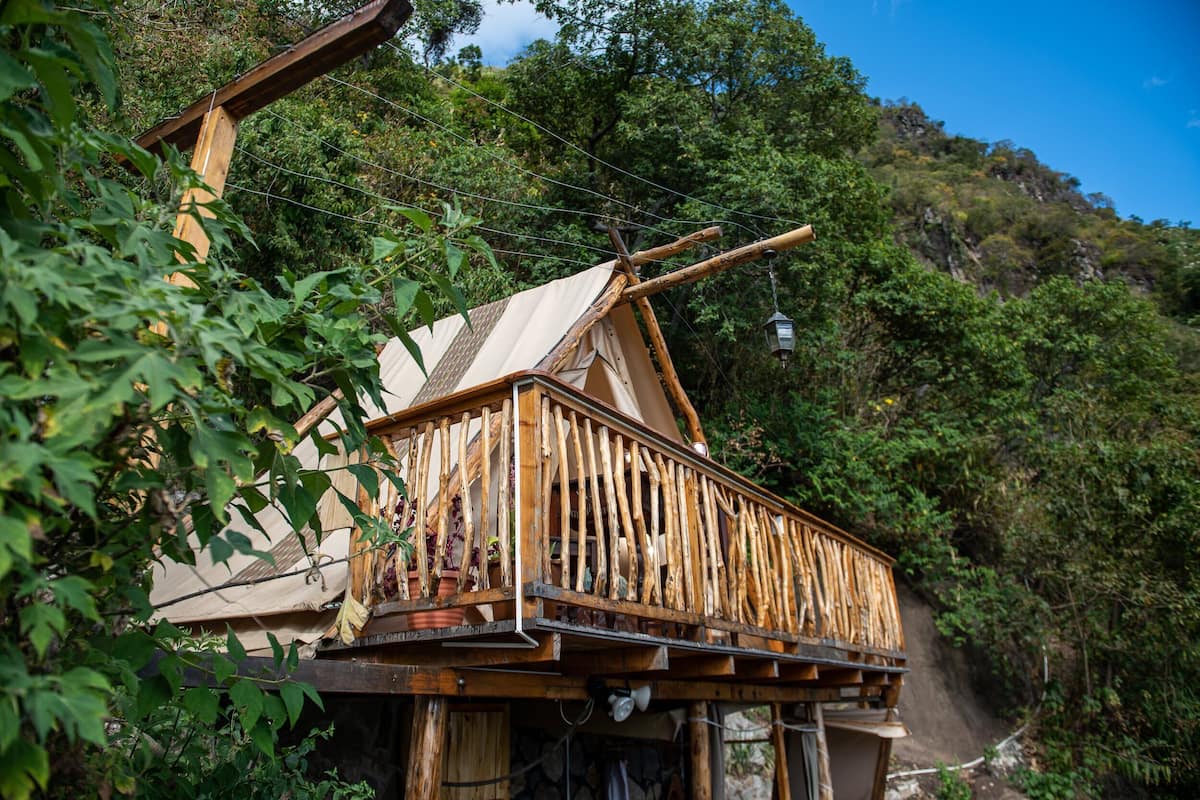 A wooden cabin with a balcony, surrounded by lush greenery and a steep hillside.