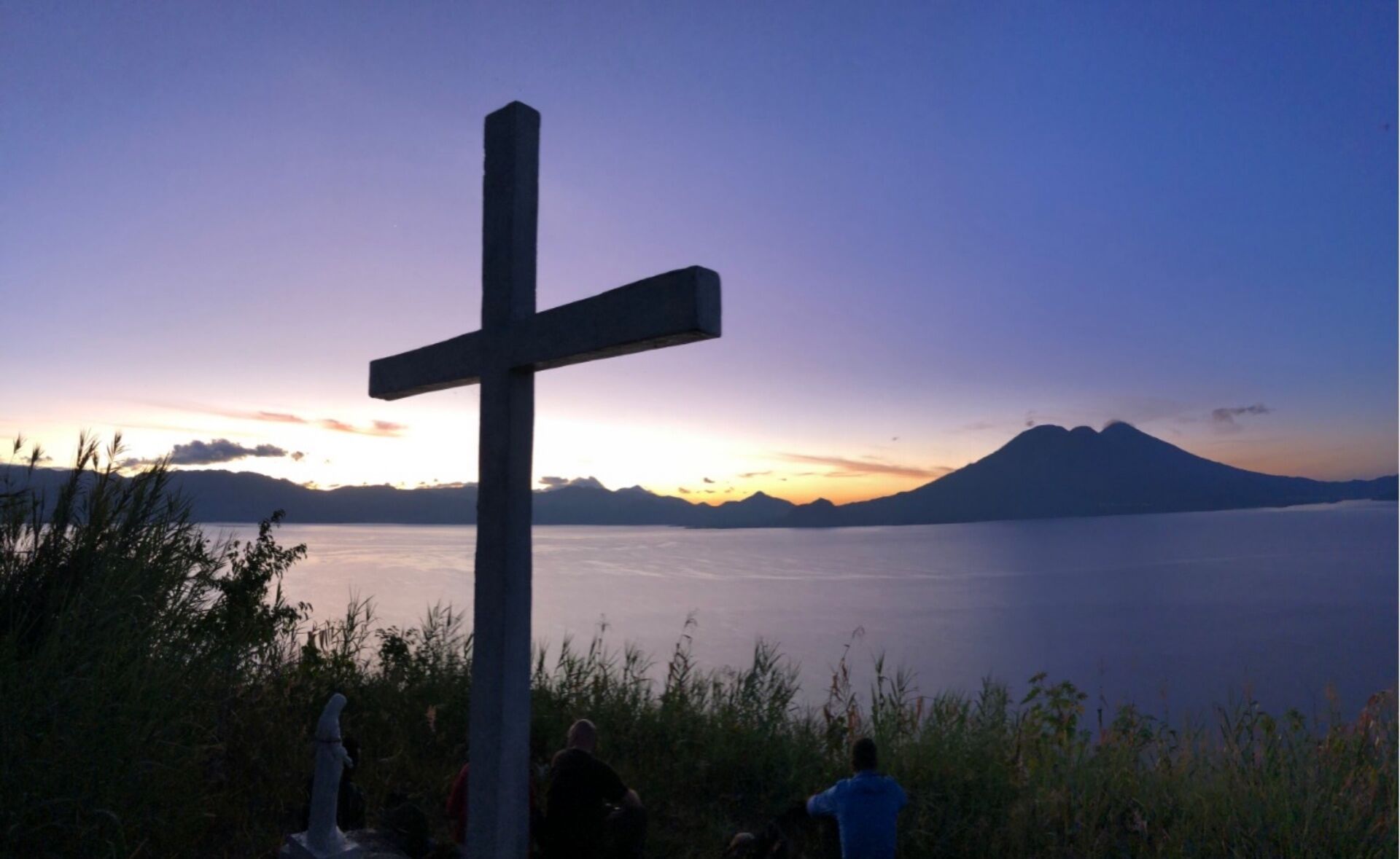 Ein Kreuz mit Blick auf Berge und ein Gewässer bei Sonnenuntergang.