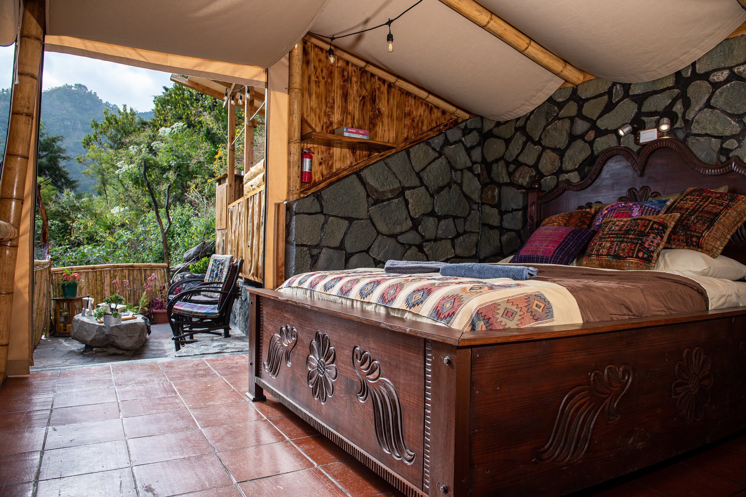 Un dormitorio con pared de piedra, cama de madera con manta colorida, una silla y vistas a zonas verdes al exterior.