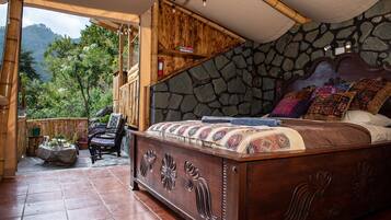 Un dormitorio con pared de piedra, cama de madera con manta colorida, una silla y vistas a zonas verdes al exterior.
