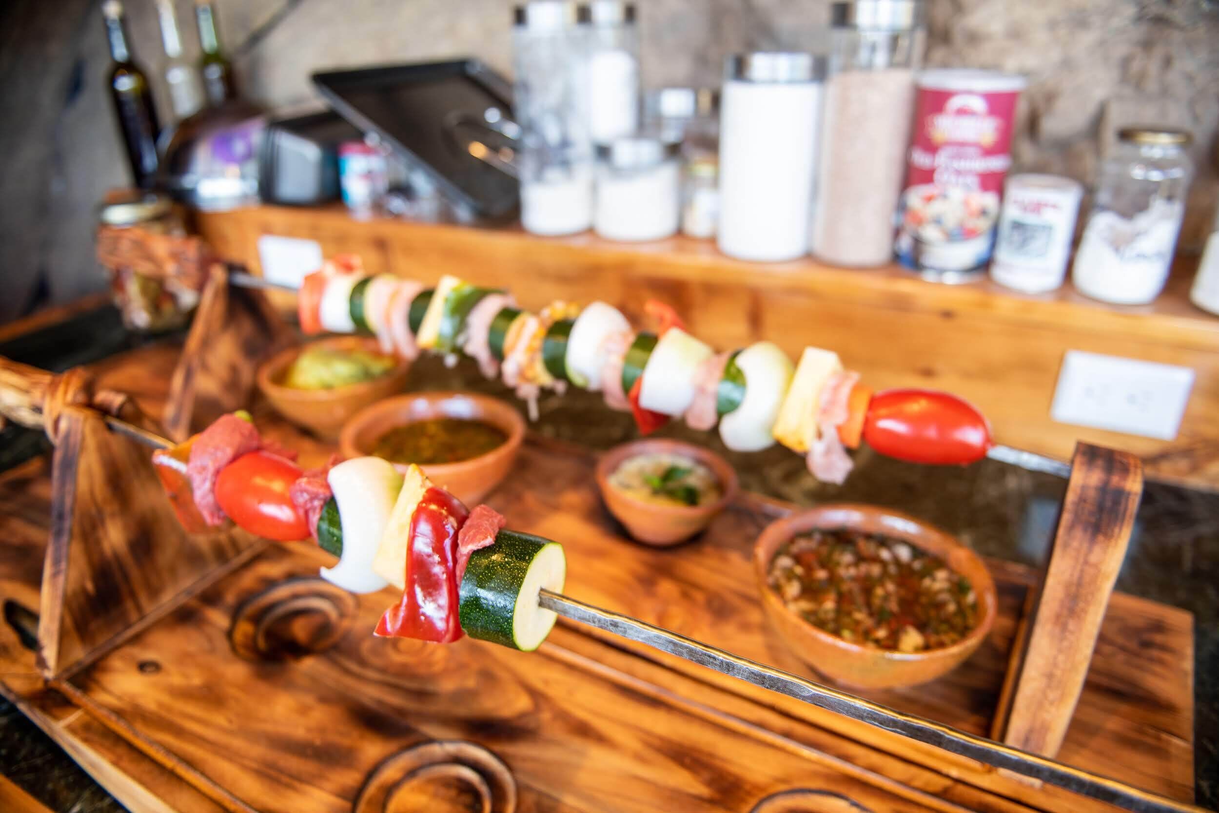 Una bandeja de madera con brochetas de verduras y carnes, acompañadas de recipientes pequeños con salsas.
