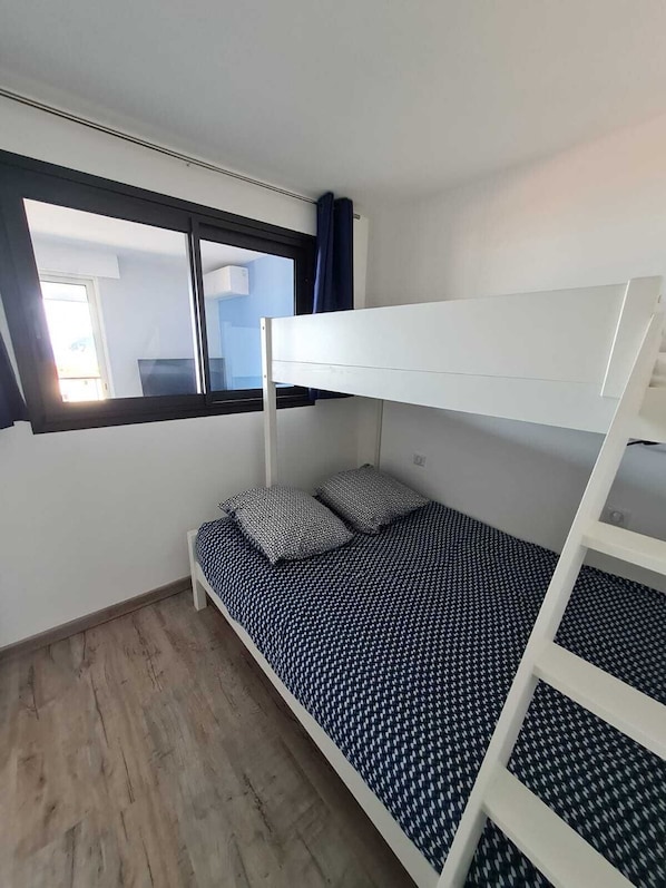 Quarto com beliche, janela, piso de madeira e edredom estampado.