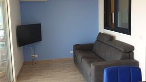 Una sala de estar con un sofá gris, un sillón azul, un televisor montado y una ventana con persianas.