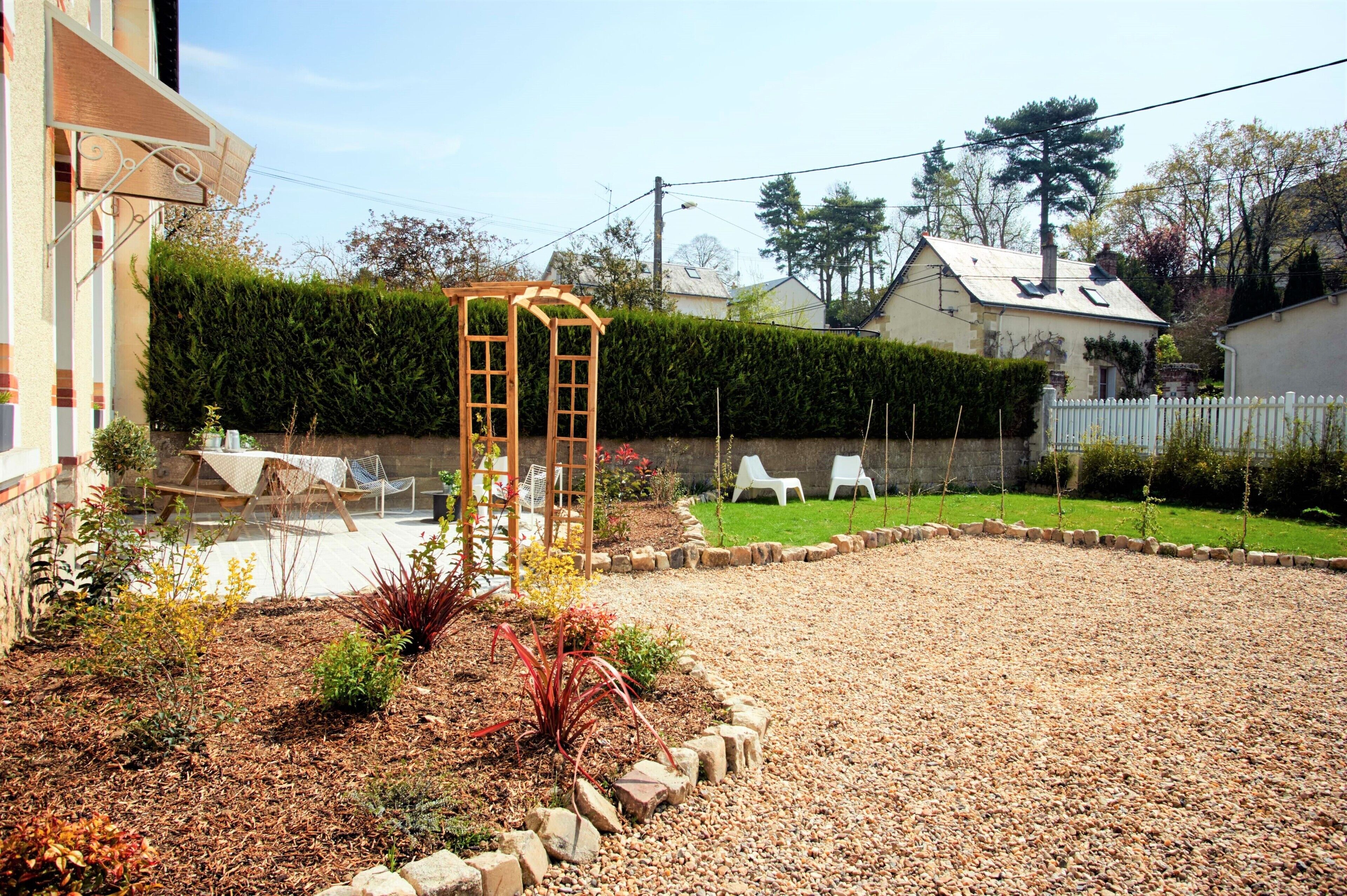 Ein Garten mit einer Holzpergola, weißen Lounge-Stühlen, einer Kiesfläche und einer ordentlich geschnittenen Hecke.