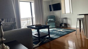 A living room with a sofa, a coffee table, a TV mounted on the wall, a chair, and a window with blinds.