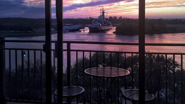 A balcony with two tables and chairs, overlooking a river with a ship and a sunset.
