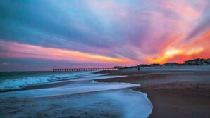 A beach with a pier extending into the water, a clear sky, and a few people walking along the shore. - Water Street Retreat 08 (Wilmington)