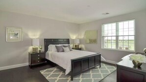 A bedroom with a large bed, two bedside tables with lamps, a window with blinds, and a small desk with a vase.