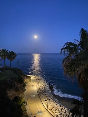 Une route côtière bordée d’une côte rocheuse, de palmiers et offrant une vue dégagée sur l’océan, sous un ciel nocturne où l’on peut apercevoir la lune.