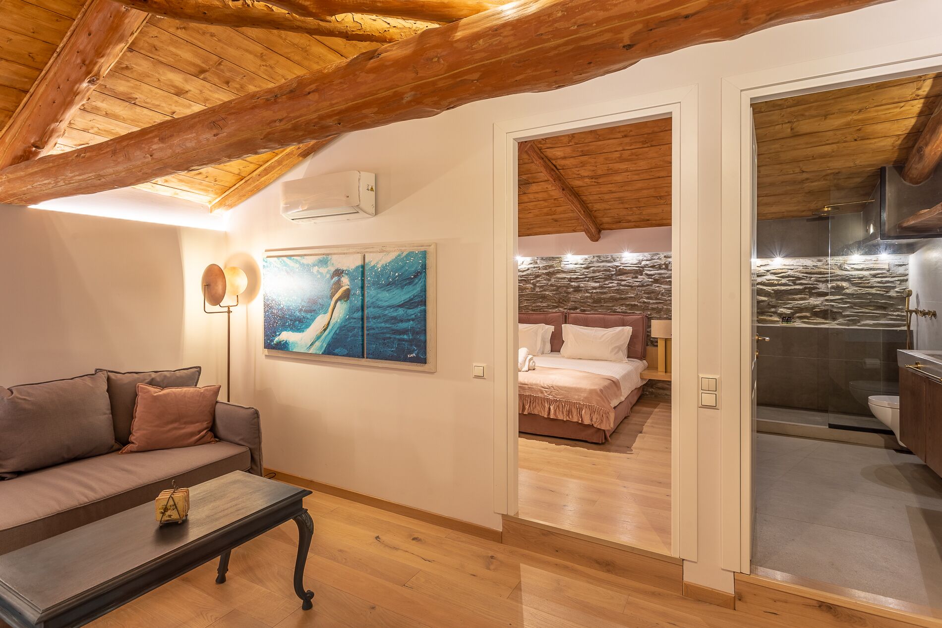 A cozy living room with a sofa, a coffee table, and a painting. There is a bedroom visible through an open door with a bed and a stone wall.