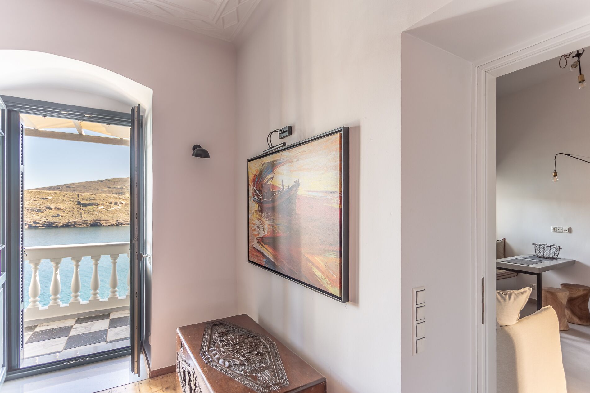 Una habitación con un amplio ventanal que ofrece vistas a un paisaje pintoresco, un cuadro colgado en la pared y una pequeña mesa de madera con intrincados grabados.