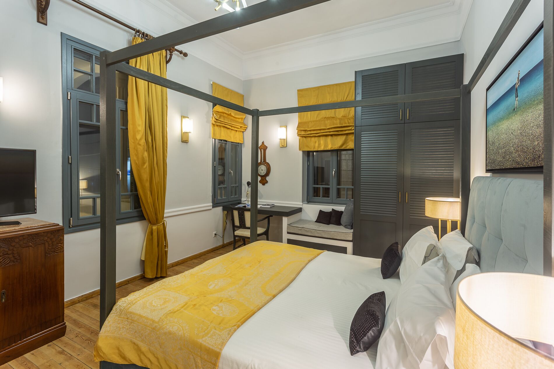 A bedroom with a four-poster bed, a desk with a chair, a small seating area, and a wardrobe.