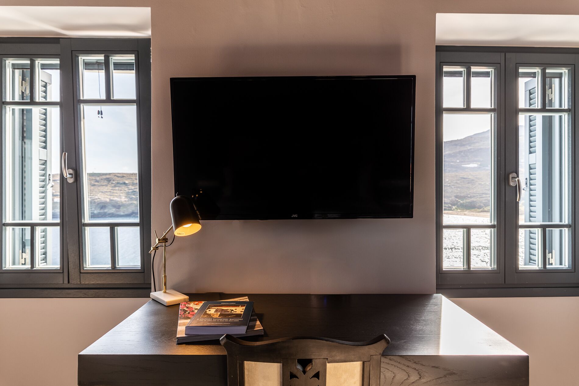 A room with a large flat-screen TV mounted on the wall, a desk with a lamp, and a chair. There are windows with blinds on either side of the TV.