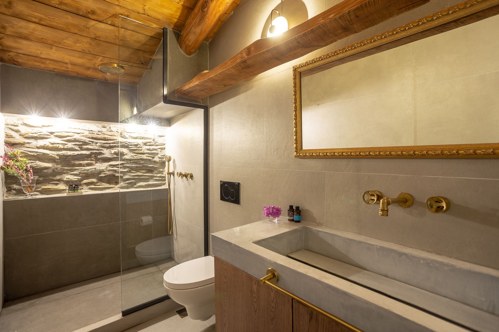 A bathroom with a stone wall, a glass-enclosed shower, a toilet, a bathtub, and a large mirror with a gold frame.