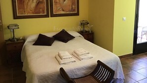 A hotel room with a bed, two paintings on the wall, a nightstand with a lamp, and a chair. - El Magnolio Rural Cottage (Santa Cruz de Pinares, Ávila)