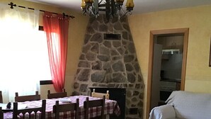 A dining area with a stone fireplace, wooden chairs, and a table set with a checkered tablecloth. - El Magnolio Rural Cottage (Santa Cruz de Pinares, Ávila)