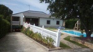 A two-story house with a front porch, a swimming pool, and a garden with a white fence. - El Magnolio Rural Cottage (Santa Cruz de Pinares, Ávila)