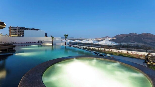 Una piscina dalla forma sinuosa, circondata da un muretto basso e un vialetto. Sullo sfondo, edifici e montagne sotto un cielo sereno.