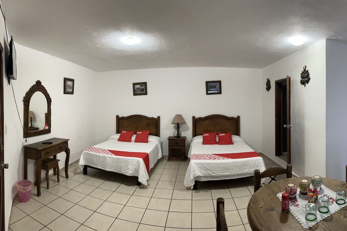 A hotel room with two beds, a wooden wardrobe, a round dining table with a lace tablecloth, and a tiled floor.