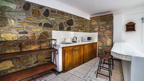 Une cuisine avec un mur en pierre, des placards en bois, un plan de travail blanc, une bouilloire, un four à micro-ondes et deux tabourets.