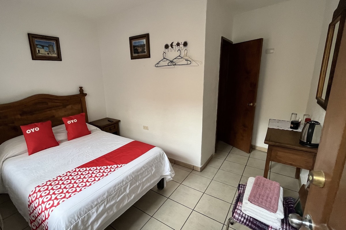 A hotel room with a wooden bed, two red pillows, a white bedspread, a small bedside table with a kettle, and a door leading to another room.