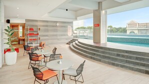 Ein moderner Poolbereich im Innenbereich mit einer Sitzecke, die runde Tische und Stühle bietet, einer Treppe, die zum Pool führt, und Blick auf das Wasser durch große Fenster.