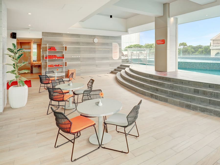 A modern indoor pool area with a seating area featuring round tables and chairs, a staircase leading to the pool, and a view of the pool through large windows.