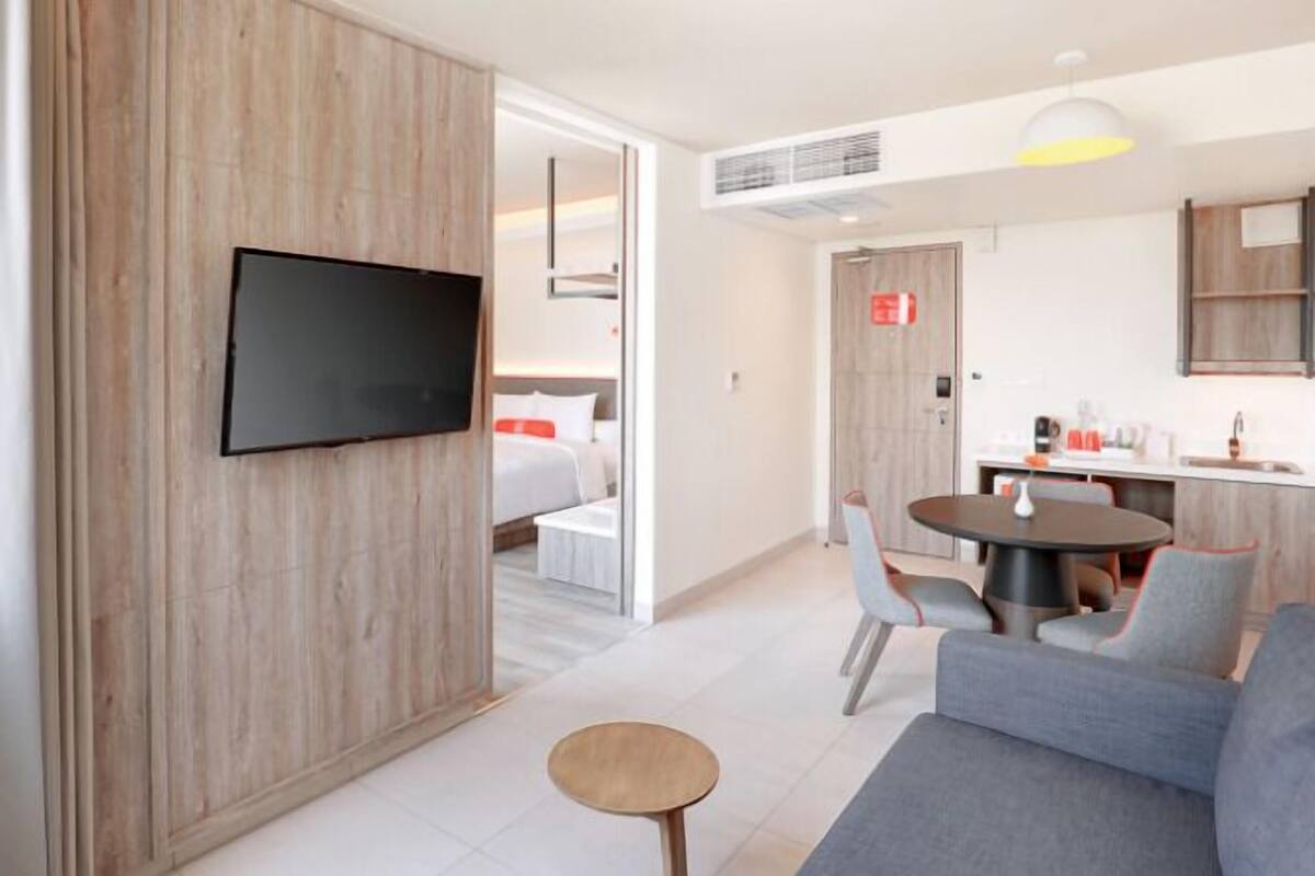 A modern hotel room with a flat-screen TV mounted on a wooden wall, a dining area with a round table and chairs, and a kitchenette with a sink and countertop.