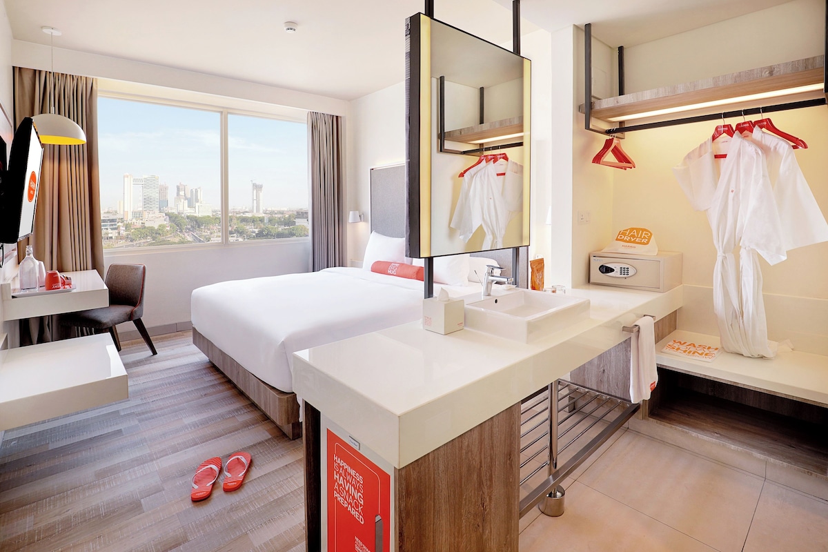 Habitación de hotel con cama, escritorio, baño con espejo y vista a la ciudad.