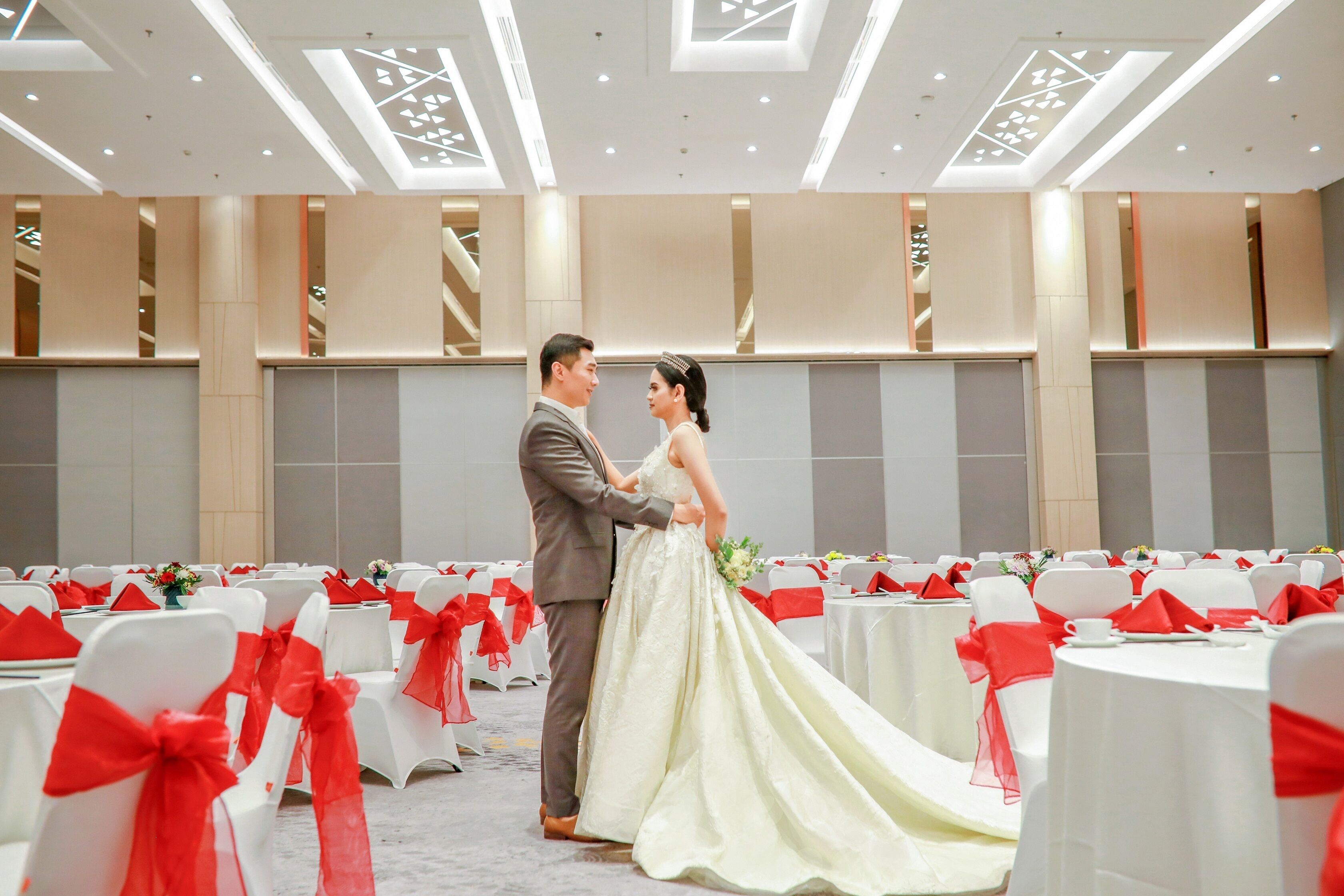 A wedding ceremony in a spacious hall with white tables and red sashes, featuring a modern ceiling design.