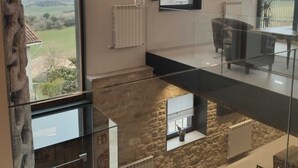 A spacious living area with a high ceiling, exposed wooden beams, a large window offering a view of the countryside, a stone wall, and a glass railing overlooking a lower level.