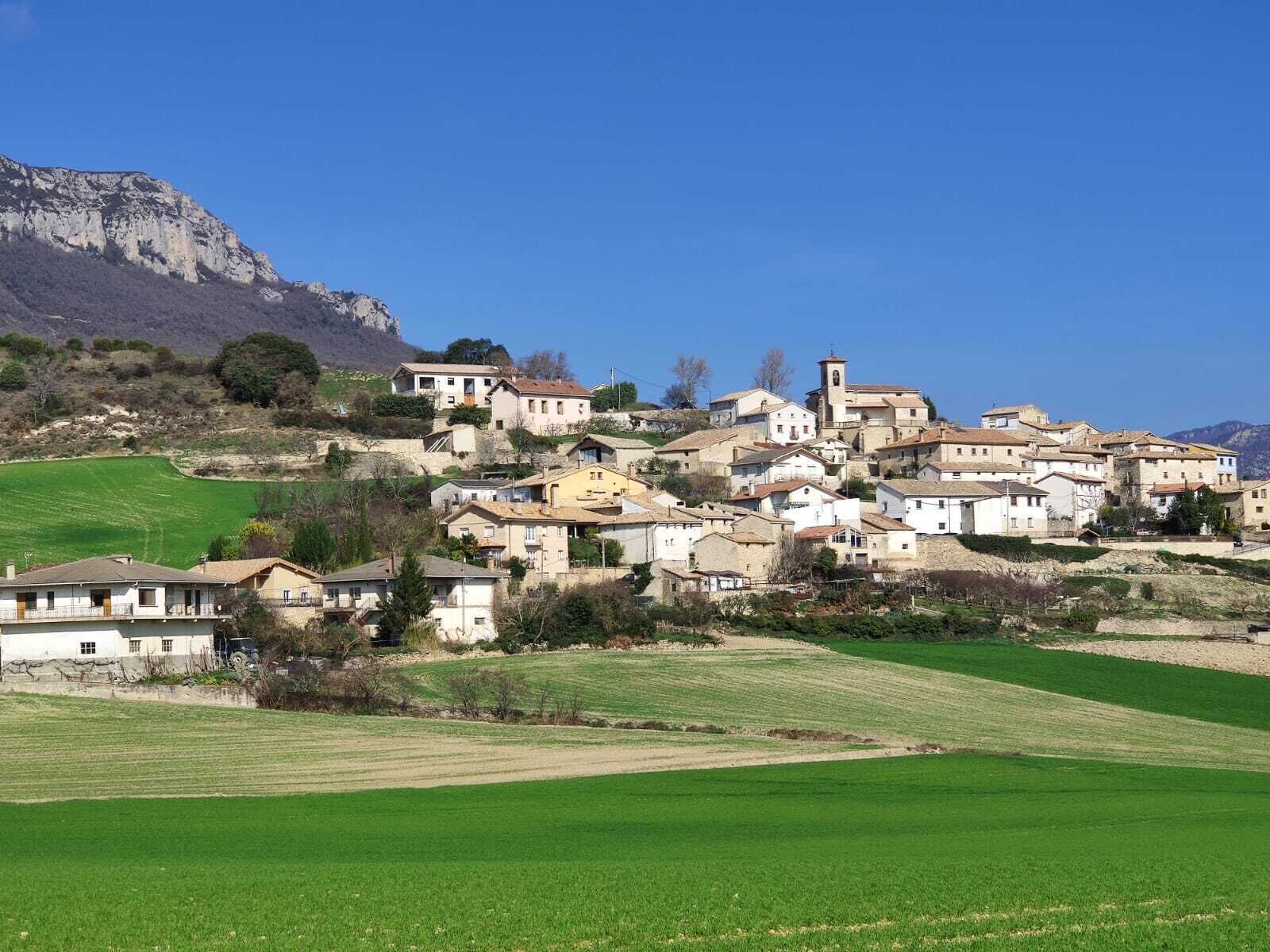 Un pueblo en una ladera con edificios de piedra tradicionales, una iglesia y una colina destacada al fondo.