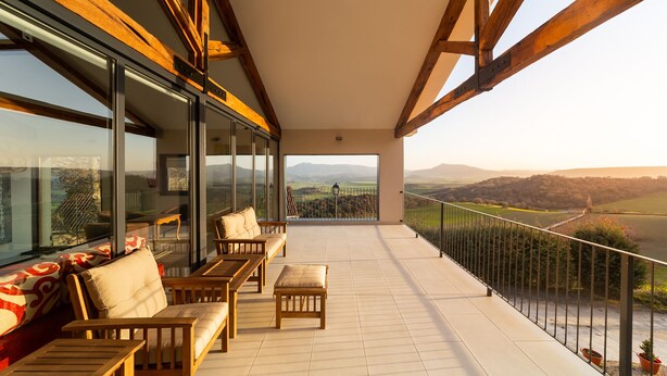 A spacious balcony with wooden furniture, large windows, and a scenic view of hills.