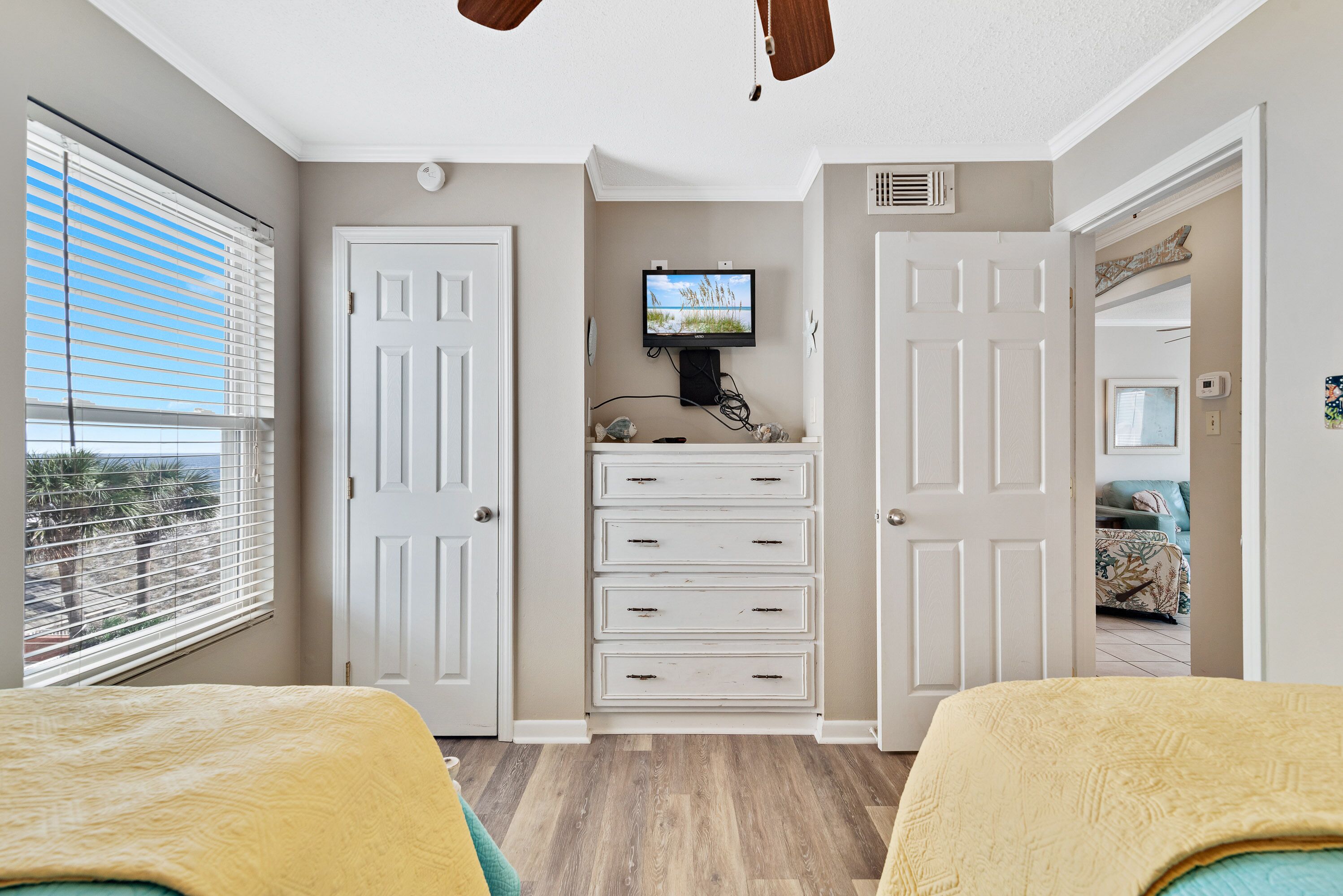 A bedroom with a TV mounted on the wall, a window with blinds, and a door leading to another room.