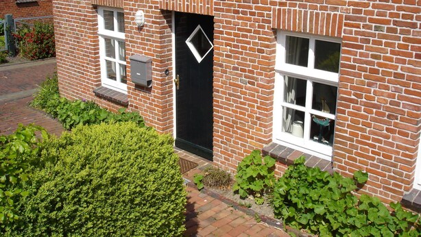 Una casa de ladrillo con puerta negra, dos ventanas y un pequeño jardín con arbustos verdes.