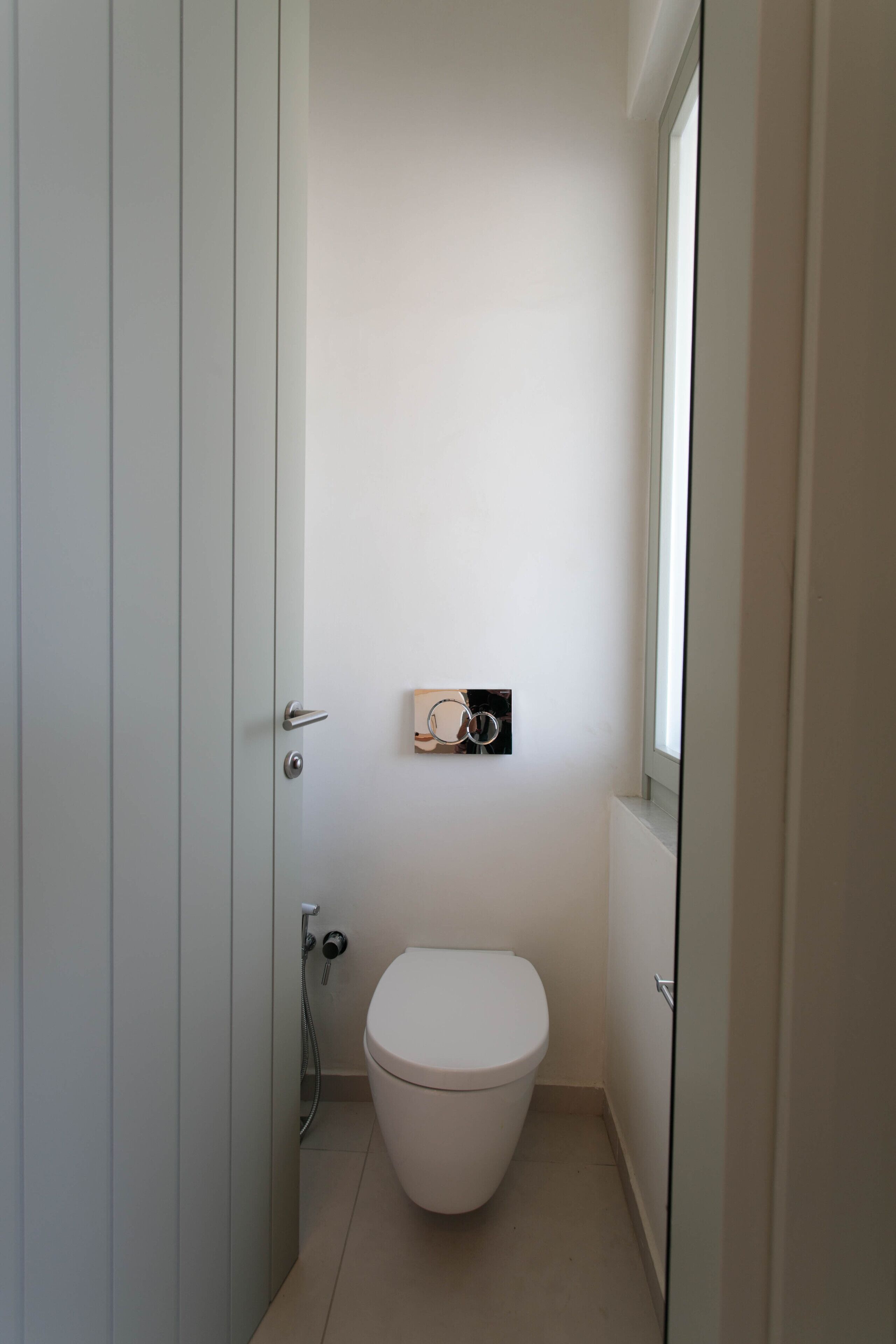 Een kleine, smalle badkamer met een wit toilet, een handdoekhouder en een raam.