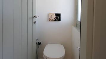 Een kleine, smalle badkamer met een wit toilet, een handdoekhouder en een raam.